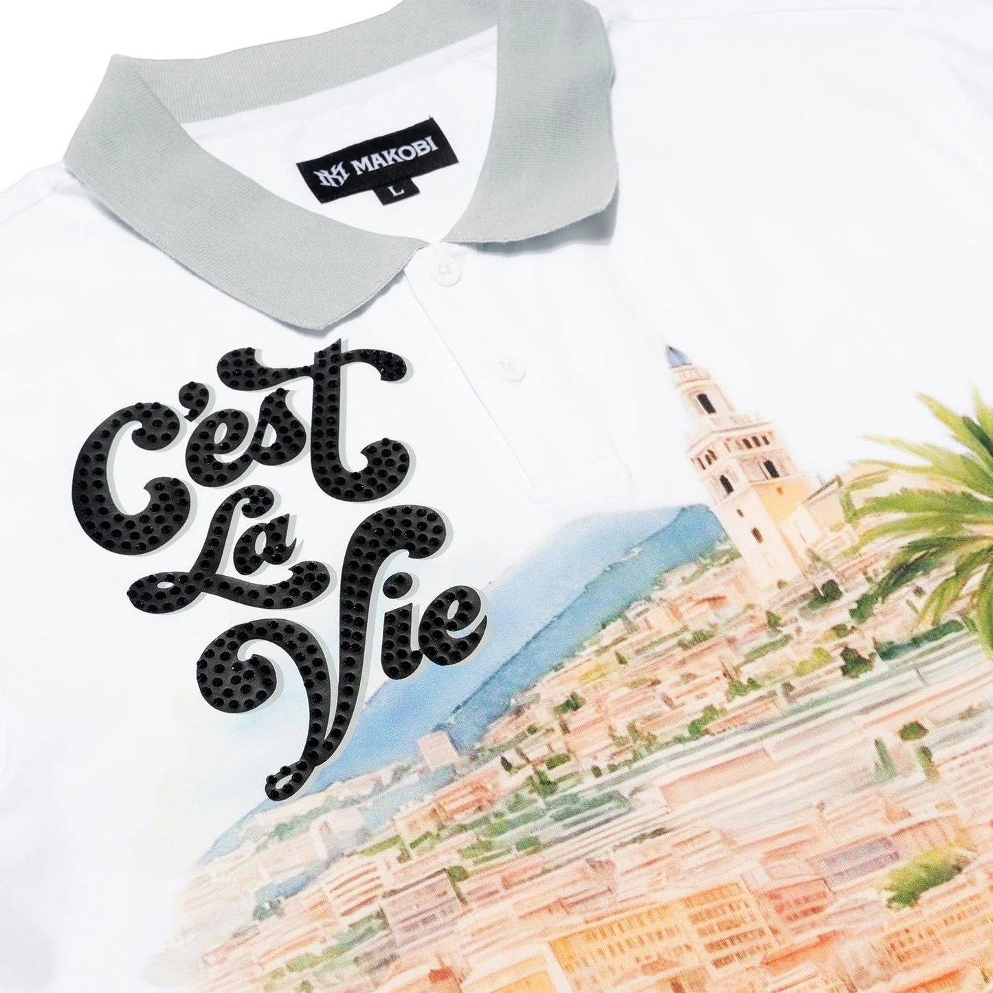 On Time Fashions white polo shirt with grey collar, featuring black rhinestone 'C'est La Vie' text and a scenic town print.